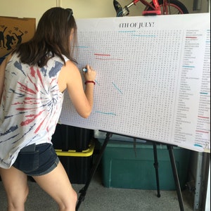 May include: A person is working on a large word search puzzle on an easel. The puzzle is titled "4TH OF JULY!" and features a list of words. The person is wearing a red, white, and blue tie-dye tank top and denim shorts. A red bicycle is visible in the background.