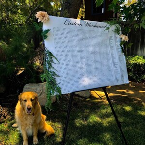 Pu&ograve; includere: Un puzzle di parole crociate personalizzato bianco su un cavalletto con un cane dorato seduto di fronte. Il puzzle di parole crociate &egrave; decorato con vegetazione e fiori.