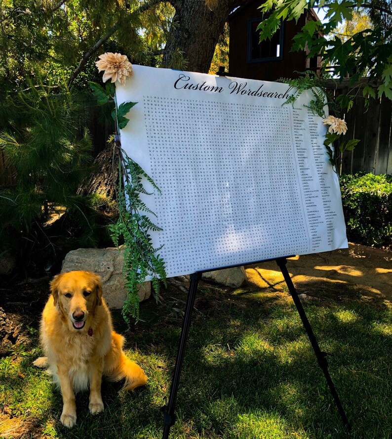 Pu&ograve; includere: Un puzzle di parole crociate personalizzato bianco su un cavalletto con un cane dorato seduto di fronte. Il puzzle di parole crociate &egrave; decorato con vegetazione e fiori.