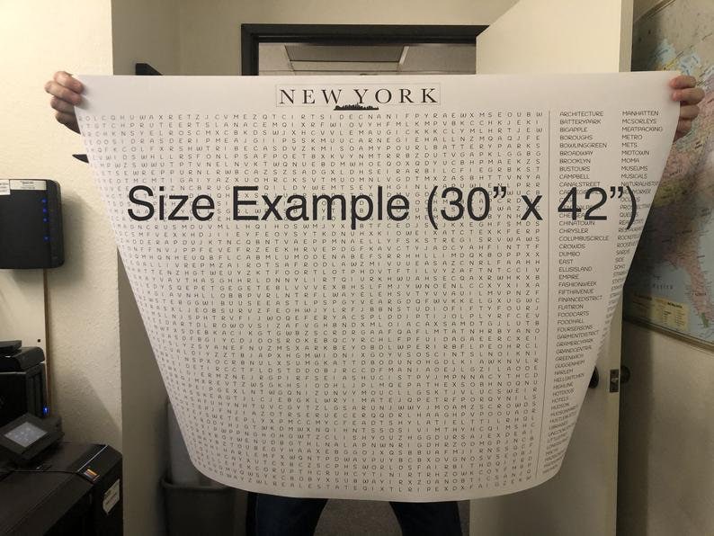May include: A black and white word search puzzle featuring the word "NEW YORK" at the top. The puzzle is printed on a white sheet of paper and is being held by a person's hands. The text "Size Example (30" x 42")" is printed in black at the bottom of the puzzle.