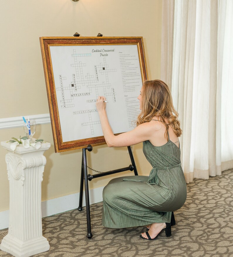 May include: A woman in a green dress is filling out a cocktail crossword puzzle on an easel. The crossword puzzle is titled "Cocktail Crossword Puzzle".