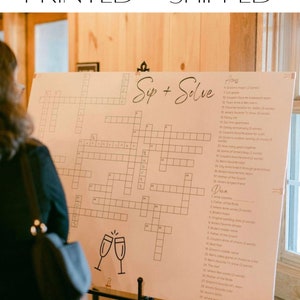 May include: A crossword puzzle game with the title "Sip + Solve" printed on a light brown background. The crossword puzzle is filled with clues related to a wedding. The clues are written in a cursive font. The crossword puzzle is on a black easel.