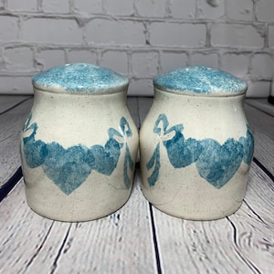 May include: A pair of ceramic salt and pepper shakers. The shakers are off-white with blue speckled lids and a blue heart and ribbon design around the base. The shakers are displayed on a wooden surface.