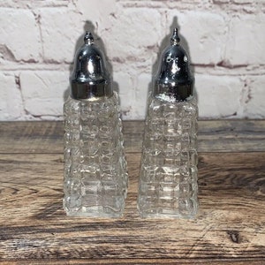 May include: Two clear glass salt and pepper shakers with silver-colored metal tops. The glass bodies have a textured, square pattern. The shakers are displayed on a wooden surface.