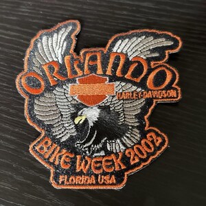 May include: An embroidered patch featuring a bald eagle with spread wings, an orange Harley-Davidson shield logo, and the text Orlando Bike Week 2002 Florida USA in orange lettering against a black background with orange borders.
