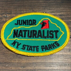 Può includere: Una toppa ovale con sfondo verde e bordo giallo. Il testo "JUNIOR NATURALIST KY STATE PARKS" è ricamato in nero. È presente anche una grafica rossa e nera di un uccello.