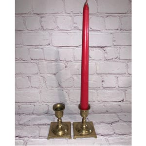 Puede incluir: Dos candelabros de latón con una vela roja. Los candelabros tienen una base cuadrada y un diseño detallado. La vela roja es alta y delgada. Sobre un fondo de ladrillos blancos.