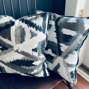 Puede incluir: Dos cojines cuadrados decorativos con un patrón geométrico en tonos grises, blancos y azul marino. Los cojines tienen un borde en contraste. Están colocados sobre una silla de madera.