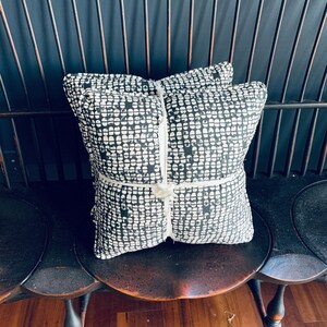May include: Two square cushions with a black and white pattern, tied together with cream string. The cushions are on a wooden chair with a dark finish, against a dark background. The cushions are approximately 40cm x 40cm.