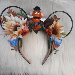 May include: A pair of black Mickey Mouse ears with a floral arrangement featuring sunflowers, blue flowers, and fall foliage. A Mr. Potato Head toy is positioned in the center of the arrangement.