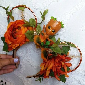 May include: Orange and green floral headband with a small brown Groot figurine. The headband is made of a copper wire frame and has a variety of orange flowers and green leaves.