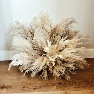 May include: A large arrangement of dried pampas grass and palm leaves in shades of beige, tan, and brown. The arrangement is placed on a wooden surface and against a white wall, creating a natural, bohemian aesthetic.