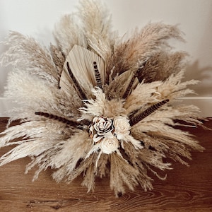 May include: A large arrangement of dried pampas grass in shades of beige and brown, with three white faux roses in the center.