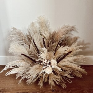May include: A dried floral arrangement featuring pampas grass, feathers, and dried roses in shades of beige and brown. The arrangement is displayed on a wooden floor.