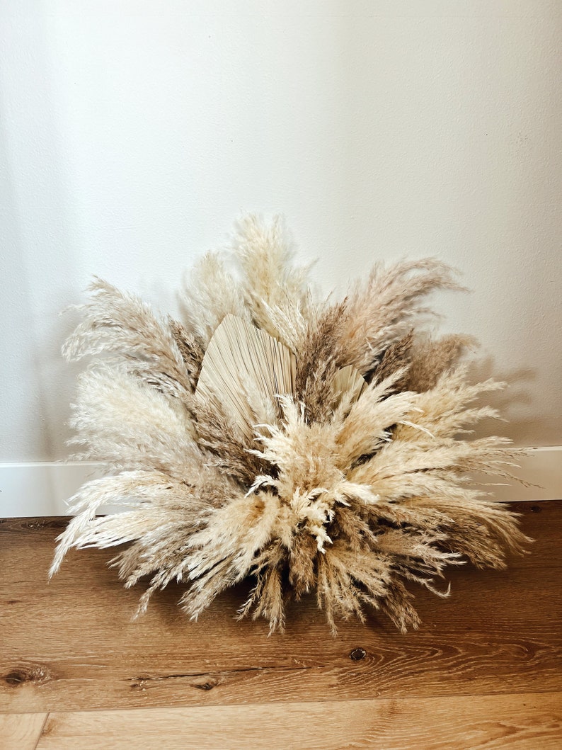 May include: A large arrangement of dried pampas grass and palm leaves in shades of beige, tan, and brown. The arrangement is placed on a wooden surface and against a white wall, creating a natural, bohemian aesthetic.