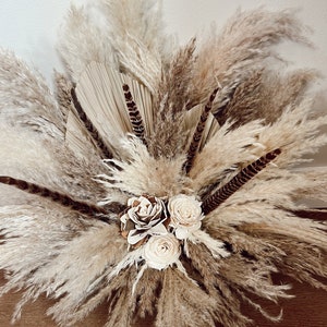 May include: A bouquet of dried pampas grass, feathers, and dried flowers in shades of beige and brown. The bouquet is arranged in a vase and is sitting on a wooden surface.
