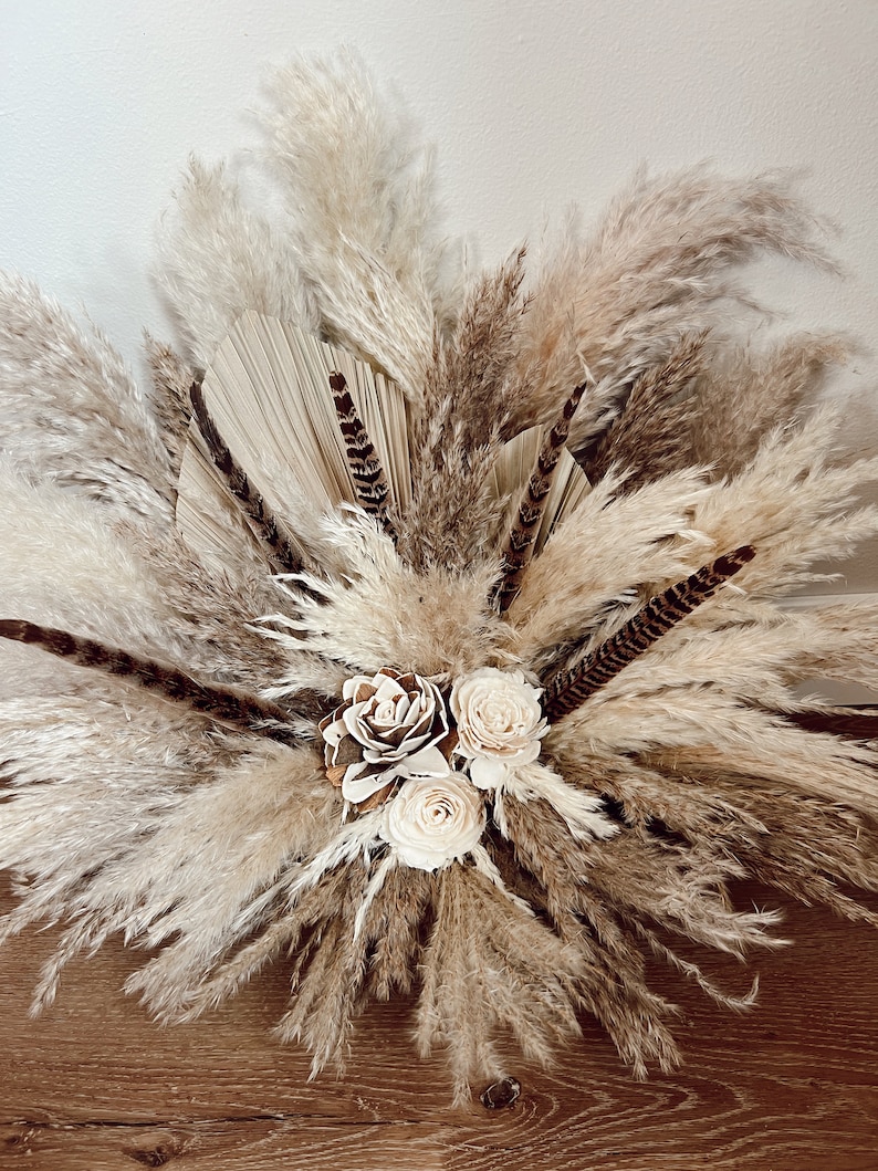 May include: A bouquet of dried pampas grass, feathers, and dried flowers in shades of beige and brown. The bouquet is arranged in a vase and is sitting on a wooden surface.