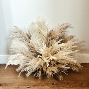 May include: A large arrangement of dried pampas grass and palm leaves in shades of beige, tan, and brown. The arrangement is set against a white wall and rests on a wooden floor. The feathery plumes create a full, textured display.
