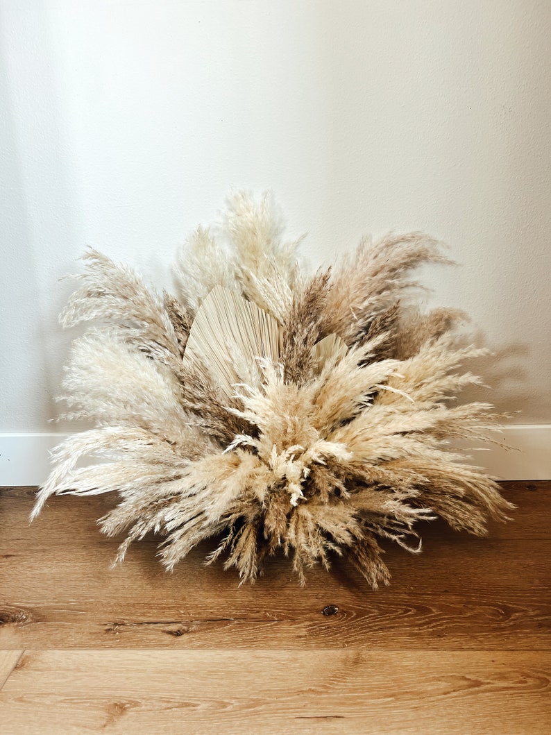 May include: A large arrangement of dried pampas grass and palm leaves in shades of beige, tan, and brown. The arrangement is set against a white wall and rests on a wooden floor. The feathery plumes create a full, textured display.