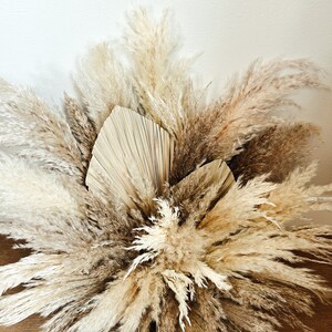 May include: A close-up of a dried floral arrangement featuring pampas grass and palm leaves in shades of beige, cream, and brown. The arrangement is set against a white wall and a wooden surface.