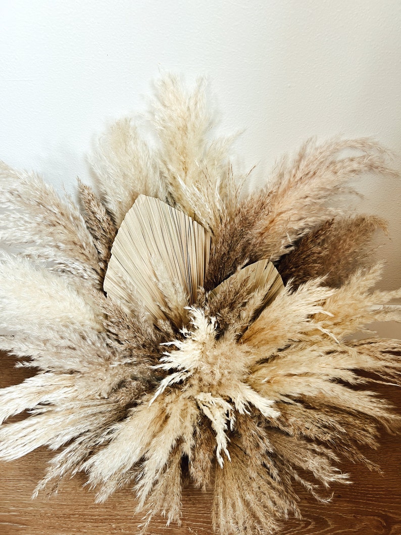 May include: A close-up of a dried floral arrangement featuring pampas grass and palm leaves in shades of beige, cream, and brown. The arrangement is set against a white wall and a wooden surface.