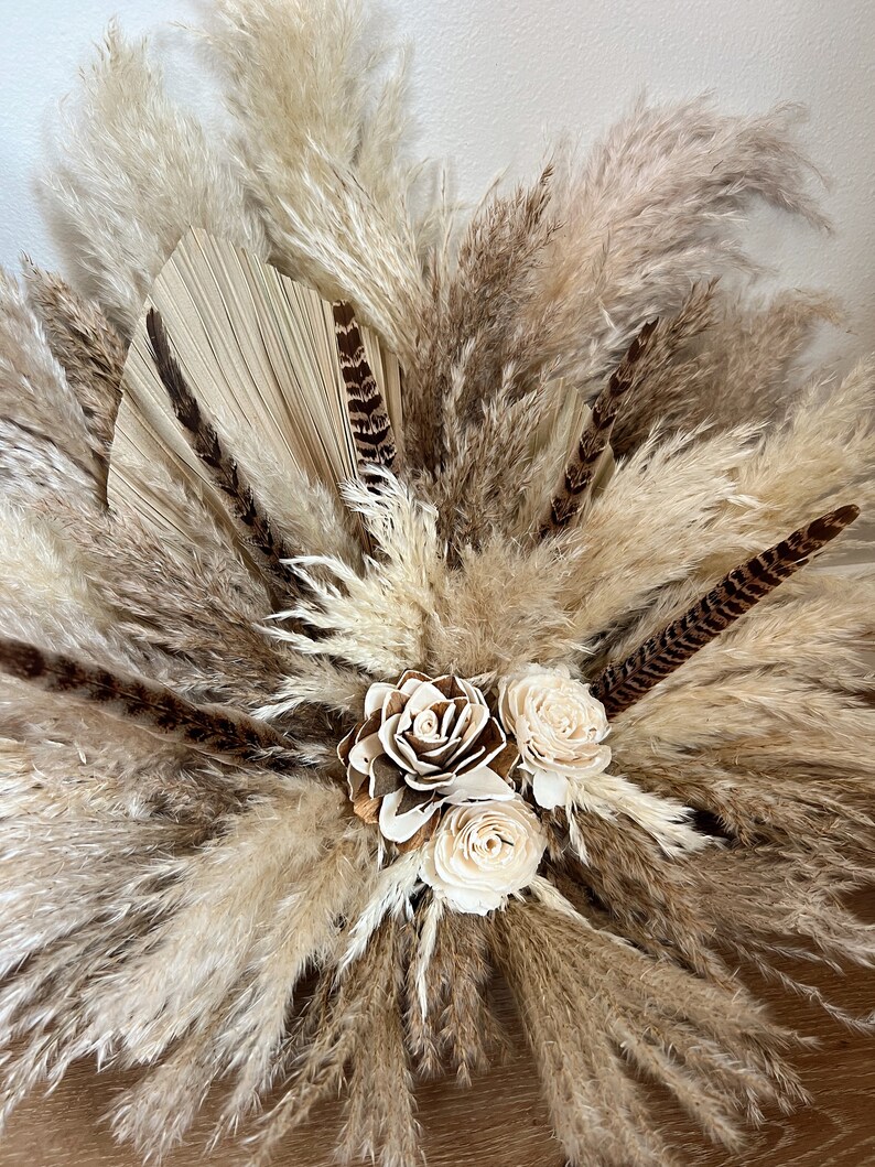 May include: A dried floral arrangement featuring pampas grass, dried palm leaves, and white dried roses. The arrangement is in a natural, earthy color palette.