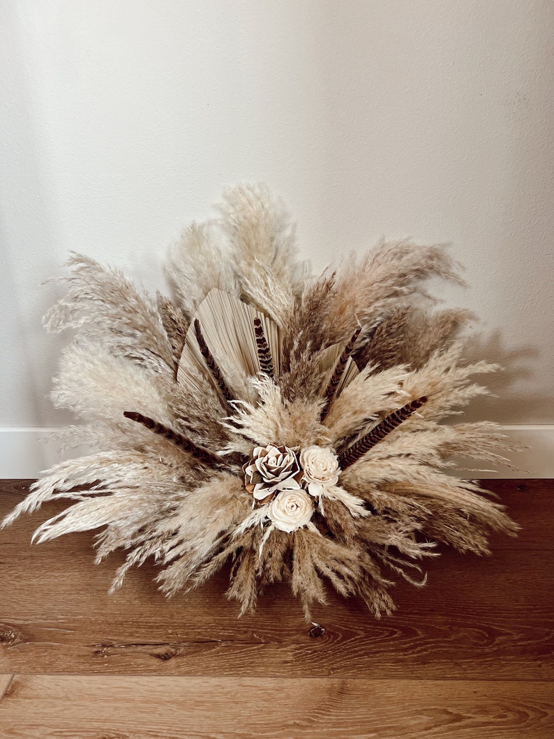 May include: A dried floral arrangement featuring pampas grass, dried palm leaves, and white faux roses. The arrangement is in a neutral color palette and has a boho aesthetic.