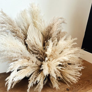 May include: A large arrangement of dried pampas grass and a dried palm leaf. The feathery plumes range in color from cream to light brown, creating a natural, neutral aesthetic. The arrangement is placed on a wooden floor against a white wall.