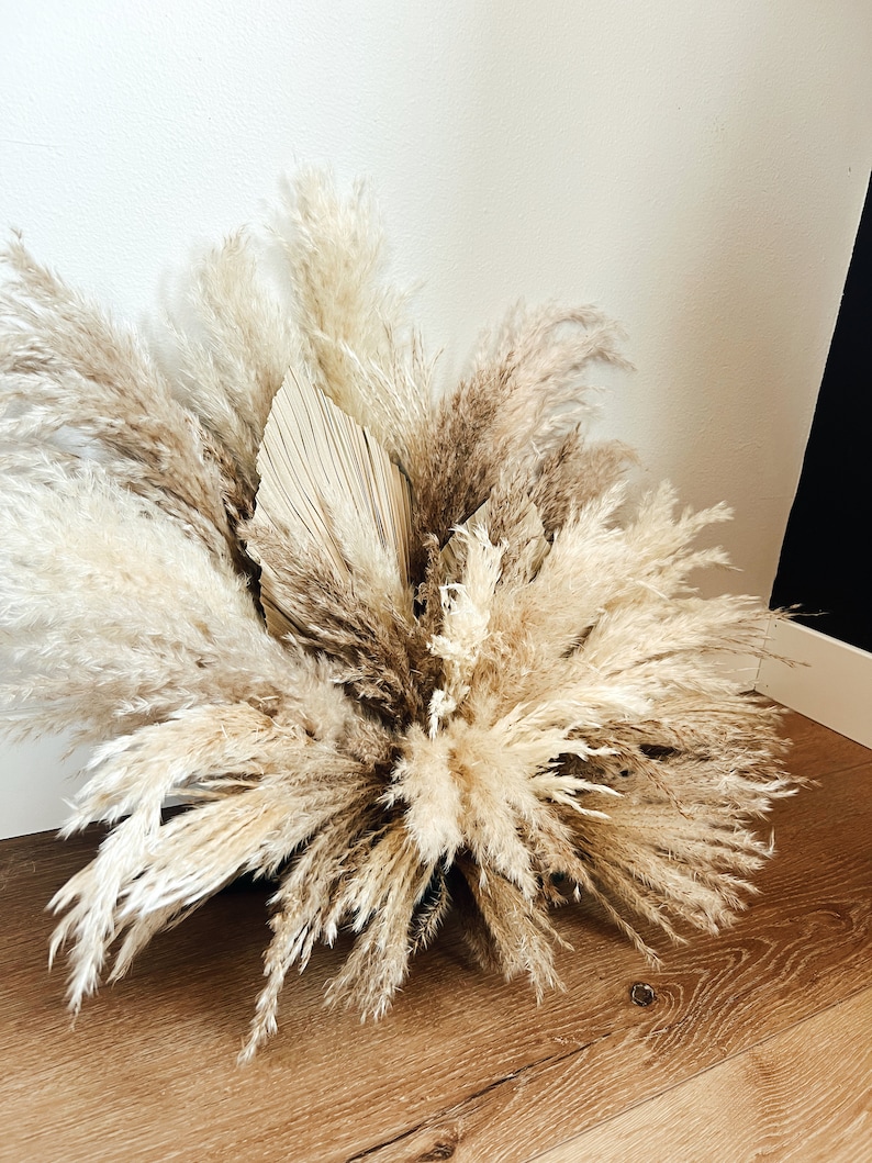 May include: A large arrangement of dried pampas grass and a dried palm leaf. The feathery plumes range in color from cream to light brown, creating a natural, neutral aesthetic. The arrangement is placed on a wooden floor against a white wall.