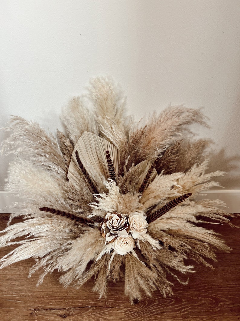 May include: A large arrangement of dried pampas grass in shades of beige and brown, with three white faux roses in the center.