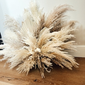 May include: A large arrangement of dried pampas grass and palm leaves in shades of beige, cream, and brown. The feathery plumes are arranged in a full, textured display, perfect for home decor or event styling.