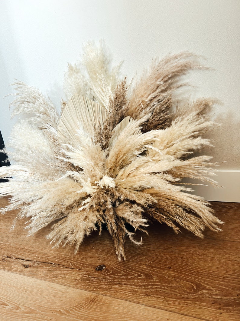 May include: A large arrangement of dried pampas grass and palm leaves in shades of beige, cream, and brown. The feathery plumes are arranged in a full, textured display, perfect for home decor or event styling.