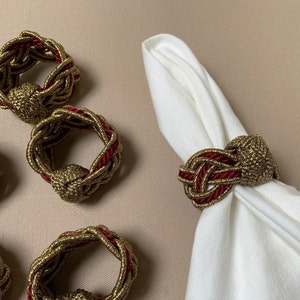 以下が含まれることがあります： 金と赤のナプキンリング5個セット。リングは、結び目が付いた編み紐で作られています。リングは白い表面の上に置かれています。