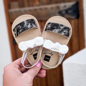 May include: A pair of baby sandals with tan soles and black and white patterned straps. Each sandal has a white bow and a strap with a velcro closure. The sandals are labeled "Baby" and "12CM".