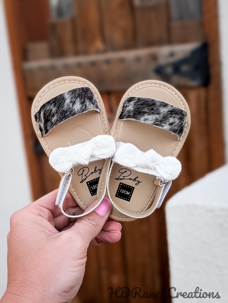May include: A pair of baby sandals with tan soles and black and white patterned straps. Each sandal has a white bow and a strap with a velcro closure. The sandals are labeled "Baby" and "12CM".