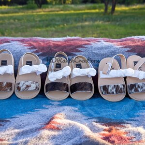 May include: Six pairs of baby sandals with cowhide accents and white bows. The sandals have tan soles and straps, with a cowhide band across the top. The sandals are arranged on a patterned blanket.