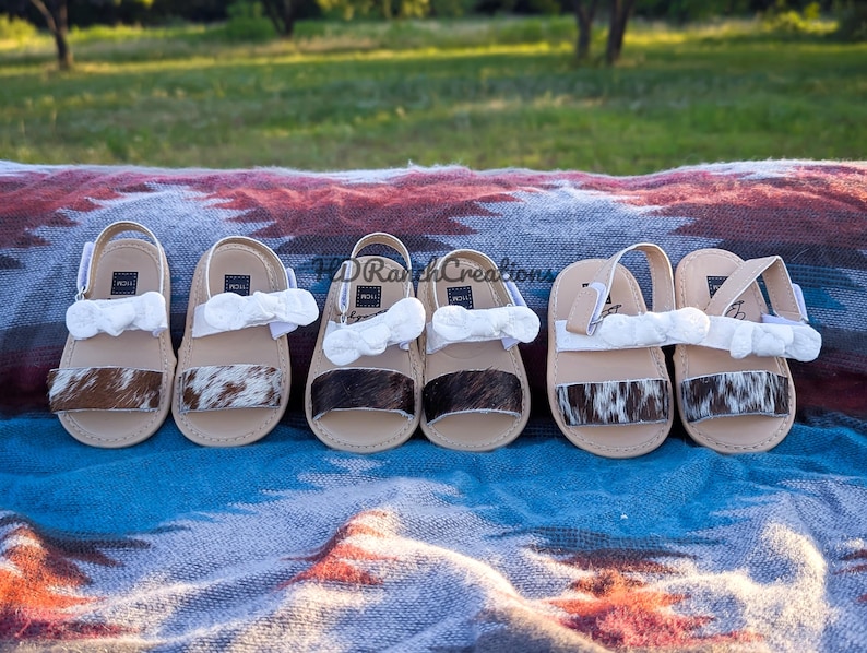 May include: Six pairs of baby sandals with cowhide accents and white bows. The sandals have tan soles and straps, with a cowhide band across the top. The sandals are arranged on a patterned blanket.