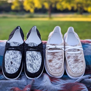 May include: Two pairs of slip-on shoes with cowhide patterns. One pair is black with white and gray cowhide, the other is tan with brown and white cowhide. Both pairs have white soles and white interiors.