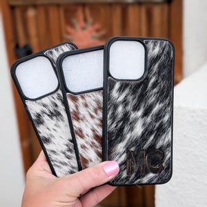 May include: Three phone cases with black edges and cowhide patterns in black and white, brown and white, and white and gray. The case on the right has the initials "MC" engraved. The cases are held in front of a wooden door.