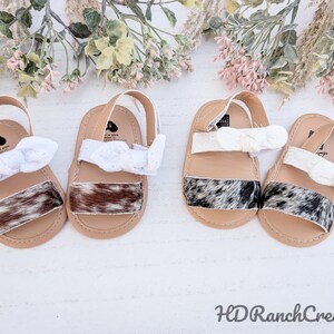 May include: Four pairs of baby sandals with tan soles, cowhide straps, and white bow accents. The sandals have a strap that goes around the ankle. The cowhide straps have a mix of brown, black, and white patterns. The background has floral accents.