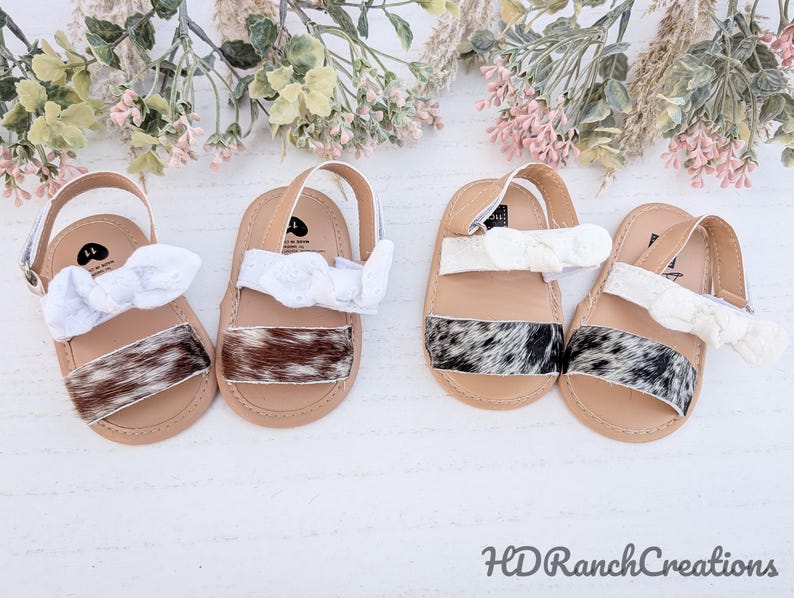 May include: Four pairs of baby sandals with tan soles, cowhide straps, and white bow accents. The sandals have a strap that goes around the ankle. The cowhide straps have a mix of brown, black, and white patterns. The background has floral accents.