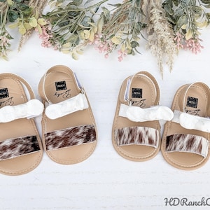 May include: Four pairs of baby sandals. Each sandal has a tan sole, a white strap, and a brown and white patterned strip across the top. The sandals have a strap around the heel. The sandals are decorated with floral accents.