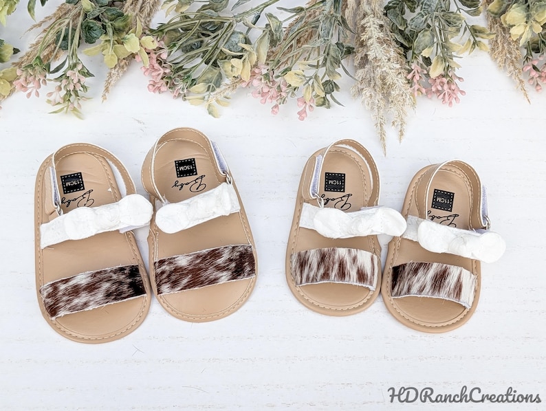 May include: Four pairs of baby sandals. Each sandal has a tan sole, a white strap, and a brown and white patterned strip across the top. The sandals have a strap around the heel. The sandals are decorated with floral accents.