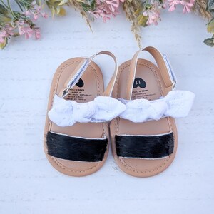 May include: A pair of baby sandals with tan soles, black accents, and white bows. The sandals have a silver strap and are size 11. The sandals are displayed on a white surface with floral accents.