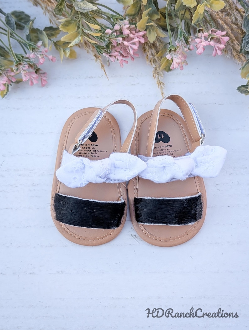 May include: A pair of baby sandals with tan soles, black accents, and white bows. The sandals have a silver strap and are size 11. The sandals are displayed on a white surface with floral accents.
