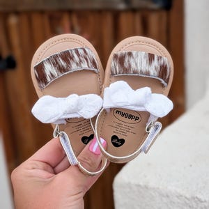 May include: A pair of baby sandals with a tan base, a brown and white cowhide accent, and a white bow. The sandals have a gold-colored trim and a small heart-shaped logo. The text "myggpp" is visible on the insole.