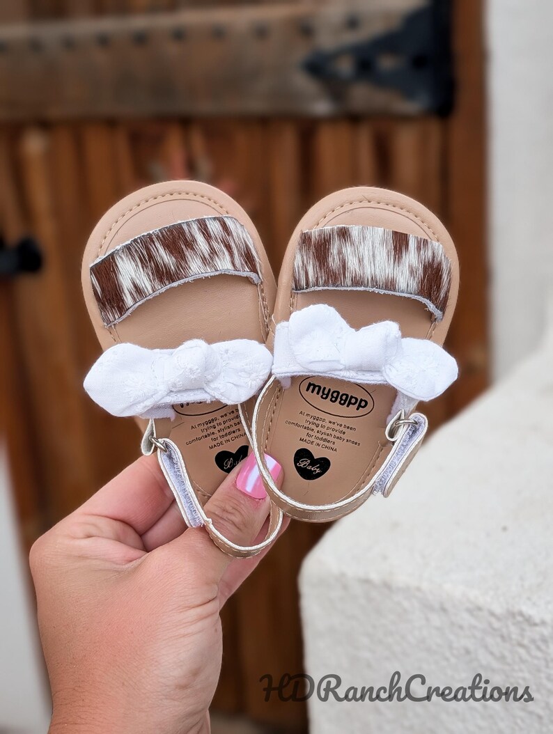 May include: A pair of baby sandals with a tan base, a brown and white cowhide accent, and a white bow. The sandals have a gold-colored trim and a small heart-shaped logo. The text "myggpp" is visible on the insole.