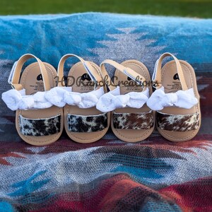 May include: Four pairs of baby sandals with tan soles, white straps, and cowhide patterned accents. Each sandal has a white fabric bow. The sandals are arranged on a patterned blanket.