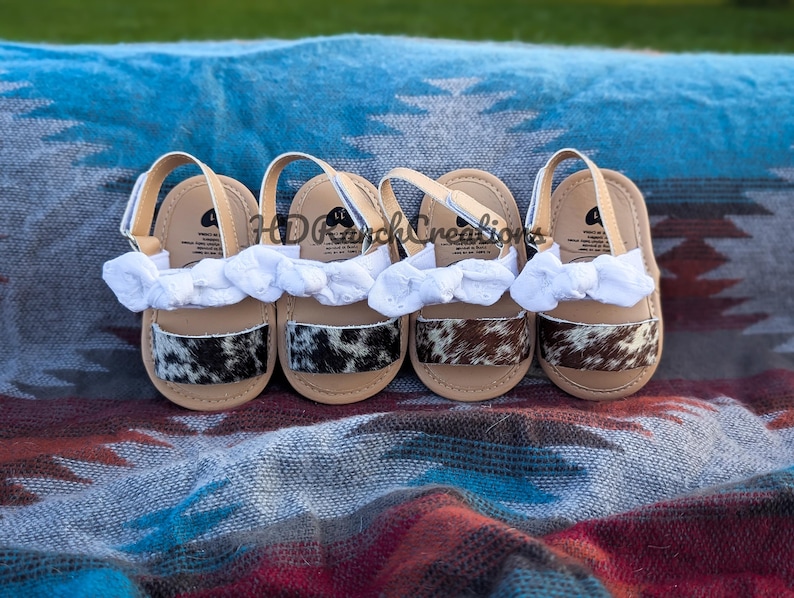 May include: Four pairs of baby sandals with tan soles, white straps, and cowhide patterned accents. Each sandal has a white fabric bow. The sandals are arranged on a patterned blanket.