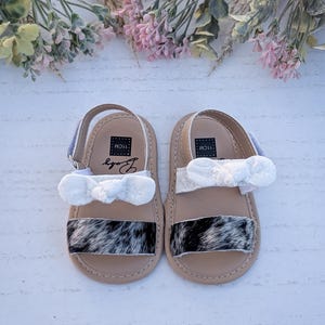 May include: A pair of baby sandals with a cowhide pattern on the toe strap. The sandals have a beige sole, white bow, and blue strap. The sandals are labeled "11cm" and "Body". The sandals are from "HDRanchCreations".